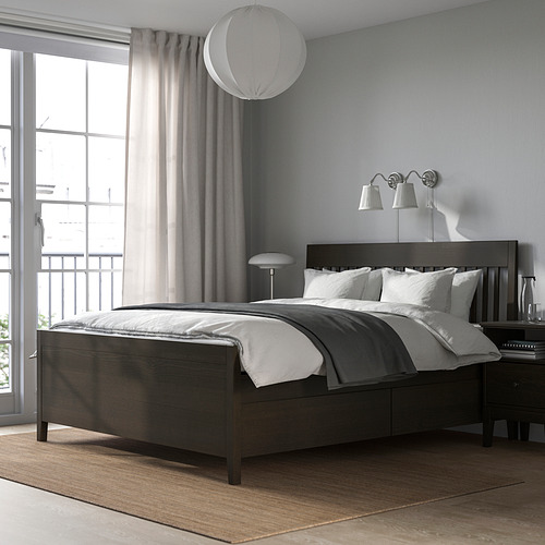Dormitorio moderno con cama de madera oscura IDANÄS con cajones de almacenamiento. Ropa de cama blanca grande, apliques de pared y una alfombra de felpa. Luz natural brillante a través de las ventanas.