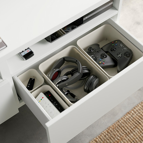 Banco de tv blanco BESTÅ con cajón abierto, almacenamiento de auriculares, controladores de juegos y controles remotos en compartimentos, y cables manejados detrás.