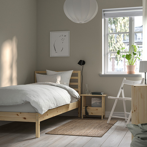 Dormitorio minimalista con cama TARVA natural, manta blanca, pared gris, mesita de noche pequeña, lámpara, planta, silla de escalera, alfombra, luz de ventana.