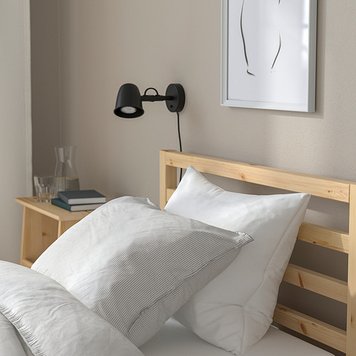 Escena de dormitorio con cama de madera TARVA, con un acogedor edredón, almohadas blancas y una lámpara negra de pared que proporciona una iluminación suave.