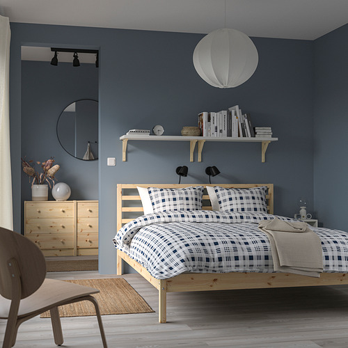 Dormitorio minimalista con cama de madera TARVA, pared azul, edredón estampado, lámpara colgante y tocador de madera. La silla marrón completa el look escandinavo.