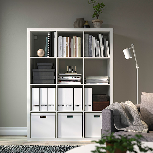 Estantería KALLAX blanca con libros, archivos y decoración. Incluye una planta en maceta y una lámpara de pie.