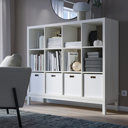 Estantería blanca KALLAX con libros, cajas y decoración, de pie junto a una silla gris.