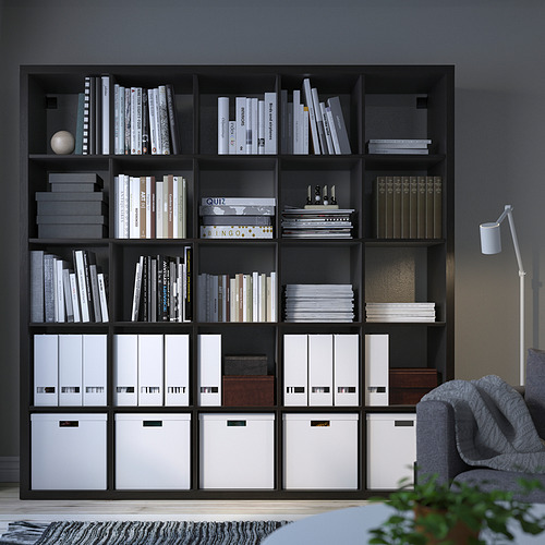 Estantería negra KALLAX, organiza libros y cajas de almacenamiento. Alto, se apoya en la pared oscura, con silla.
