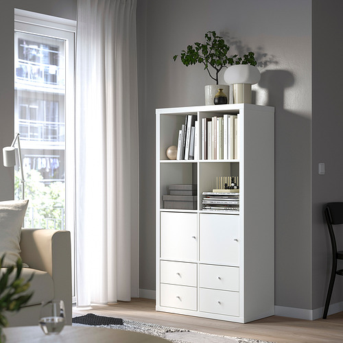 Estantería blanca KALLAX contra pared gris con libros, jarrones, plantas y ventana con cortinas.