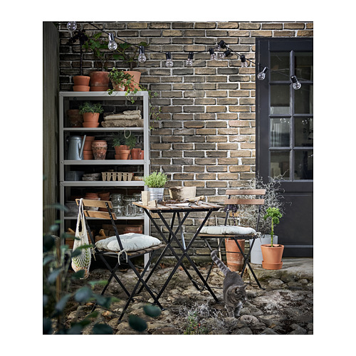 Conjunto de patio con mesa negra, sillas acolchadas y estantería para plantas. Gato cerca, fondo de pared de ladrillo con luces.