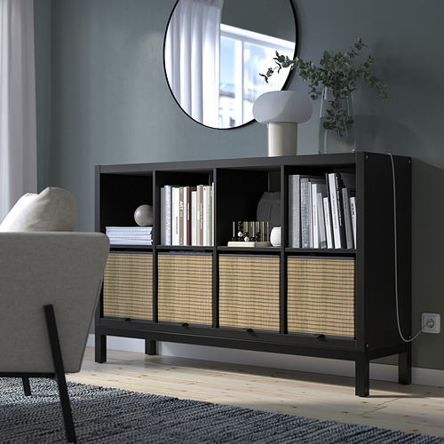 Estanterías KALLAX con cestas de mimbre, que sostienen libros y decoración, contra una pared azul. Una silla gris y un gran espejo encima realzan su diseño moderno y aireado.
