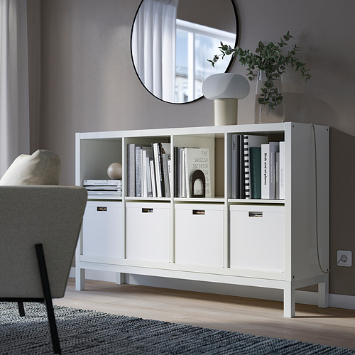 Estantería blanca KALLAX con libros y decoración cerca del sofá, base visible