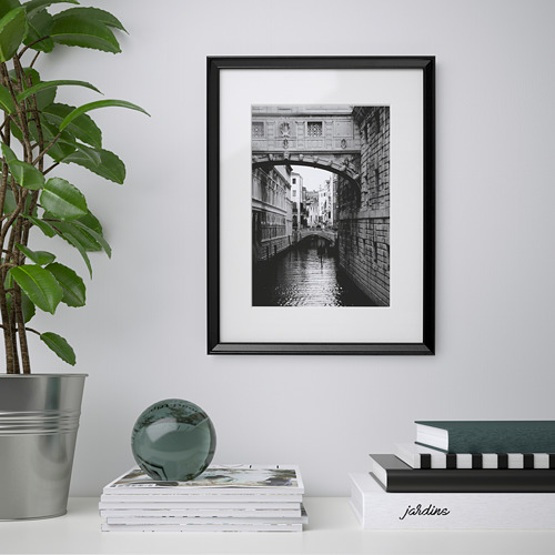 Canal de Venecia con marco negro en la pared; Debajo de él, pila de revistas con esfera de vidrio verde y planta.