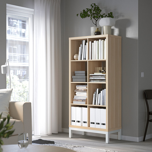 Estantería KALLAX en roble claro con cajas blancas, cerca de una ventana. Moderno, elevado, con libros y decoración.
