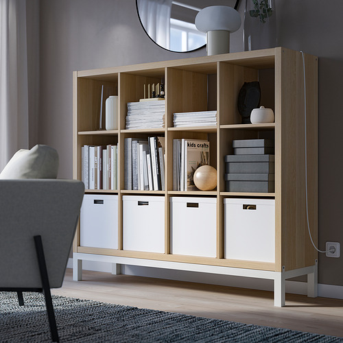 Estantería de madera clara KALLAX con silla gris, llena de cajas de almacenamiento y libros blancos y grises, enfatizando su tamaño.