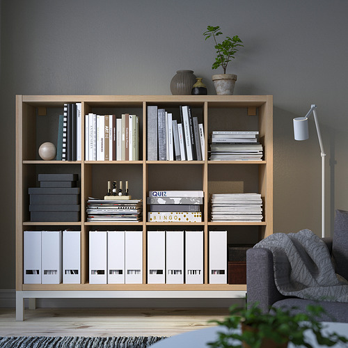 La estantería KALLAX contiene libros, juegos, archivos. Alta con múltiples compartimentos. Silla gris, lámpara blanca, planta verde cerca.
