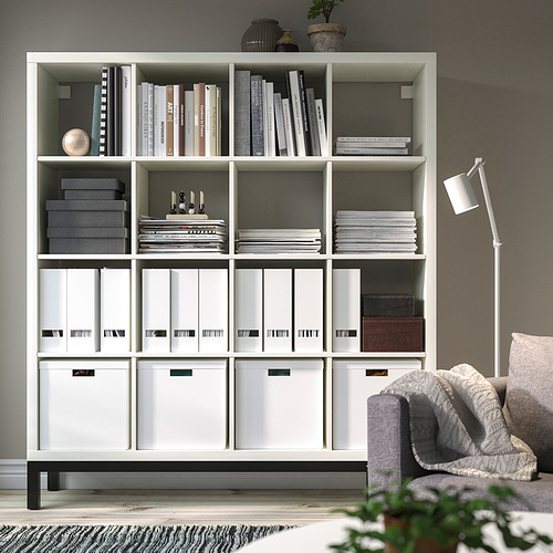 Estantería blanca KALLAX organizada con libros, cajas y decoración. Diseño alto y aireado, sirve como separador de ambientes. Sostiene varios artículos de forma ordenada.