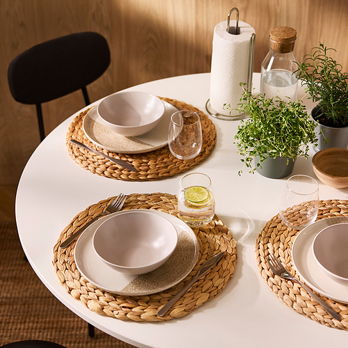Mesa de comedor redonda con manteles individuales tejidos, platos neutros y vasos. Incluye bebida de lima, hierbas y soporte para toallas de papel.