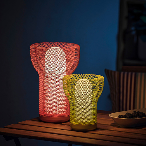 Dos lámparas SOLVINDEN ; rojo y amarillo, sobre la mesa. Diseño de malla, uso interior/exterior. El rojo es más alto, moderno y decorativo. Tazón de fruta al lado.
