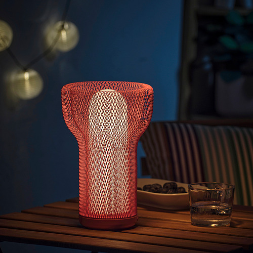 Lámpara de malla roja única en la mesa con luces, vidrio y cojines. La luz emite un brillo cálido, ideal para uso en interiores y exteriores.