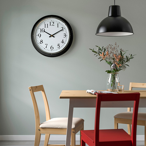 Reloj de pared BRAVUR negro sobre la mesa de comedor con flores y sillas.