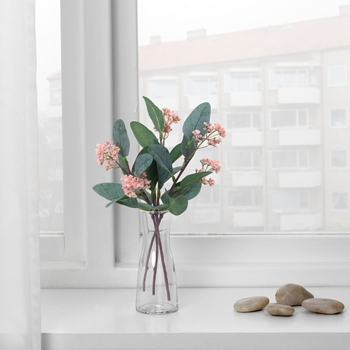 Un jarrón transparente con flores rosadas y hojas verdes se encuentra en el alféizar de una ventana blanca, junto a guijarros lisos. La luz del sol entra a raudales por la ventana.