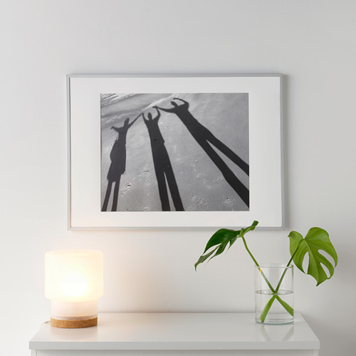 Un marco de fotos blanco y negro sombras en un estante blanco. A su lado, una pequeña lámpara encendida y un jarrón de cristal con una planta verde.
