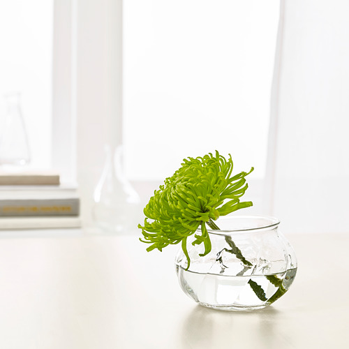 Un pequeño jarrón de vidrio, VILJESTARK, contiene una flor verde. Diseño elegante y redondo, se sienta sobre una mesa blanca.