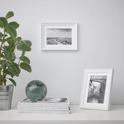 Un estante estilizado sostiene una planta, una esfera decorativa y dos marcos de fotos, uno con una imagen de un canal y otro con un paisaje, contra una pared de luz.