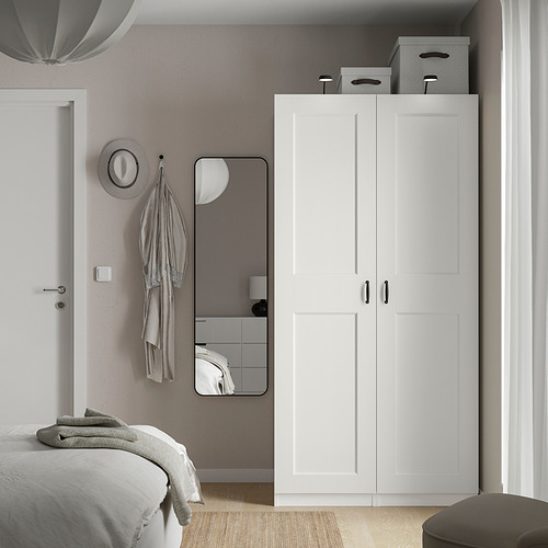 Dormitorio moderno con armario PAX blanco, cama, espejo y sombrero gris en ganchos. Diseño minimalista, líneas limpias.
