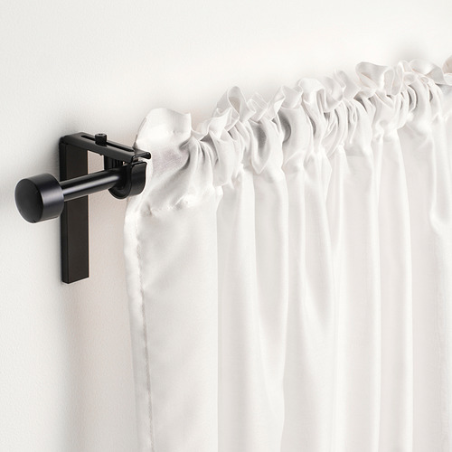 La barra de cortina negra sostiene cortinas blancas transparentes, lo que permite el paso de una luz suave. Montado en una pared gris claro.
