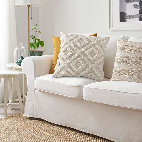 Sofá blanco con almohada grande, texturizada, color crema. Almohada amarilla también visible. Lámpara y planta en una mesita al lado del sofá. Marco de fotos en la pared.