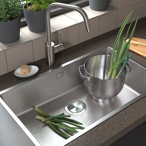 Fregadero de acero inoxidable con grifo moderno, colador lleno de cebollas verdes, hierbas frescas en ollas, estropajo, y tabla de cortar.