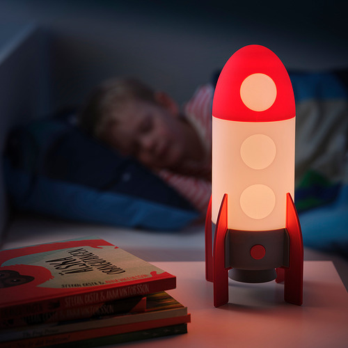 Una lámpara roja en forma de cohete en una mesita de noche, que ilumina suavemente junto a los libros infantiles.