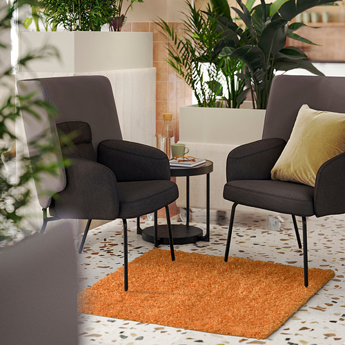 Dos sillas ÄRENDE grises con patas negras y almohadas amarillas sobre una alfombra naranja, rodeadas de plantas y una mesa auxiliar negra con una taza de cristal.