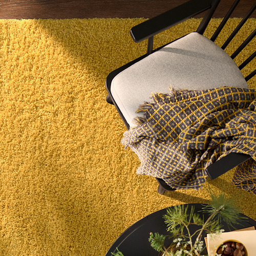 La alfombra ÄRENDE de color amarillo brillante, más suave que la silla, cubre el piso ampliamente, con una manta a cuadros oscuros y una pequeña planta cerca.