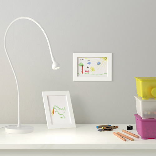 Un escritorio para niños con una lámpara flexible, dibujos en marcos blancos, lápices de colores y contenedores. La lámpara ilumina el espacio de trabajo.