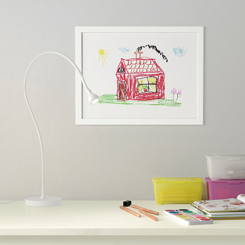 Dibujo de niños enmarcado en la pared, iluminado por una lámpara de escritorio blanca. Materiales de arte en el escritorio de abajo.