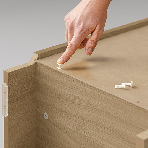 Una mano coloca una clavija blanca sobre una estantería de madera BILLY, que tiene bisagras visibles y un acabado marrón claro.