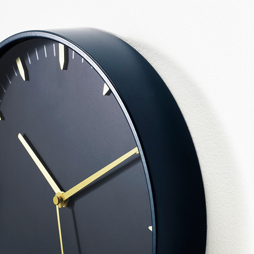 Moderno reloj de pared con manecillas doradas en esfera oscura, montado en pared blanca.