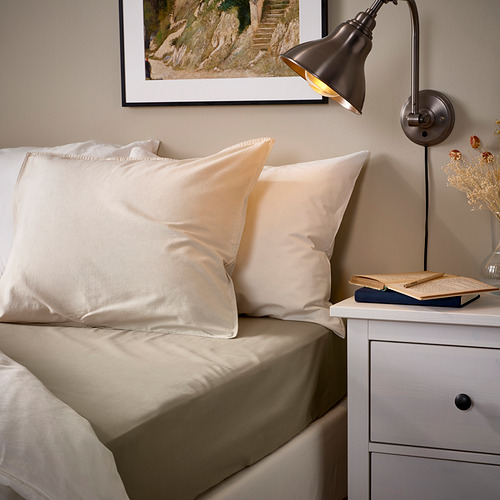 Acogedora cama con sábanas beige ULLVIDE, lámpara de pared, mesa auxiliar y arte de pared enmarcado. Las sábanas son de mezcla de algodón y lyocell, con un colchón de hasta 26 cm.