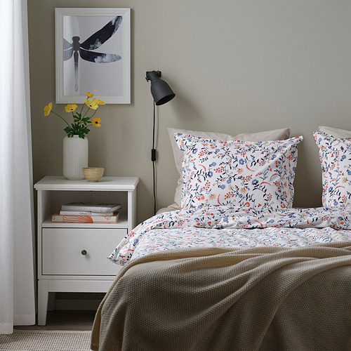 Acogedor dormitorio con juego de edredón floral azul-naranja, manta beige, mesita de noche con lámpara y flores, póster de libélula.