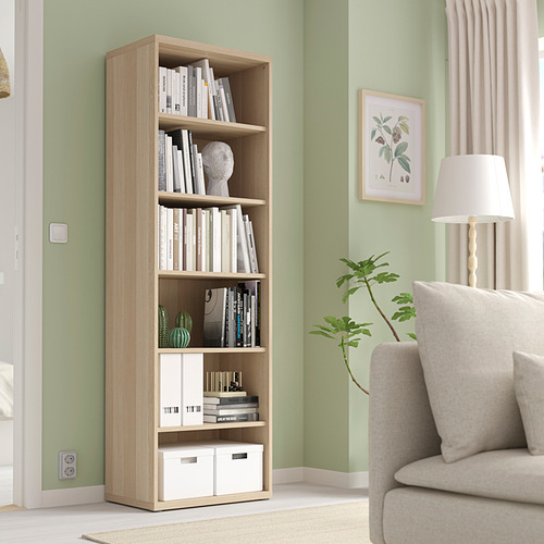 Librería de madera BESTÅ con libros y decoración, más alta que una silla y una lámpara.