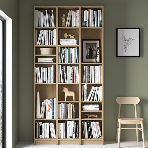 Librería BILLY de madera natural, llena de libros y decoración, contra una pared verde, más alta que una silla de madera clara.
