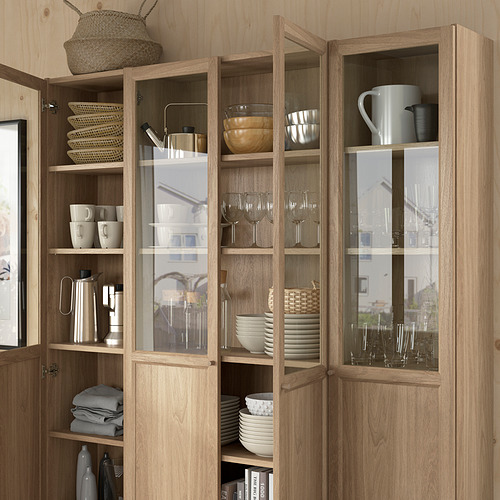Estantería de madera BILLY con frente de vidrio que muestra platos, tazas, cuencos y copas de vino en estantes ajustables.