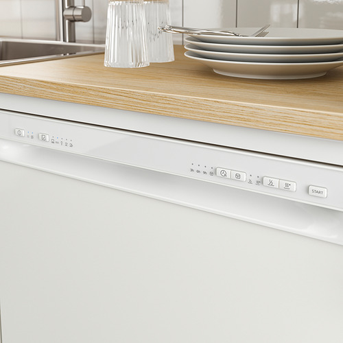 Cocina moderna con lavavajillas empotrado blanco bajo encimera de madera. Panel de control visible, cubiertos y platos apilados en el mostrador.