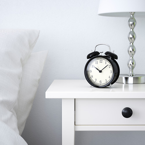 Despertador negro sobre mesita de noche blanca, que muestra la hora 12:30, junto a una almohada blanca y una lámpara.