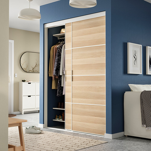 Puertas correderas de armario SKYTTA: madera clara y paneles de vidrio. Alto, se extiende desde el suelo hasta el techo, flanqueado por paredes de color azul oscuro. Diseño moderno y minimalista.