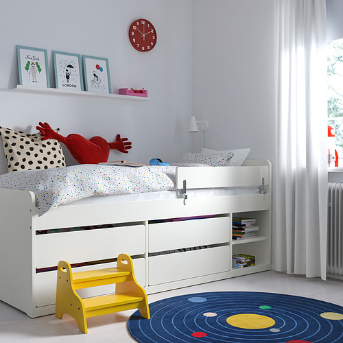 Una habitación infantil limpia con una cama SLÄKT blanca con múltiples cajones de almacenamiento. También hay un reloj rojo, coloridos estampados artísticos y un taburete amarillo.