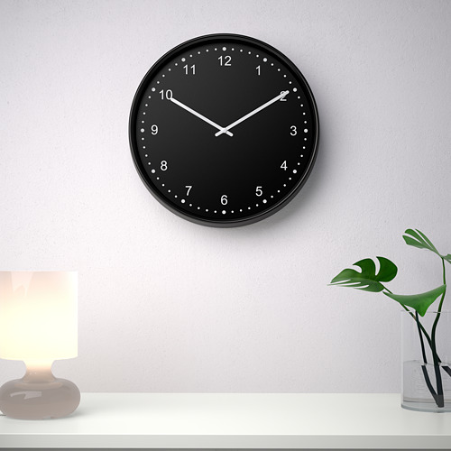 Un moderno reloj de pared negro, redondo, silencioso con números y manecillas blancos a las 10:10. Está montado sobre una estantería blanca con una lámpara y una planta.