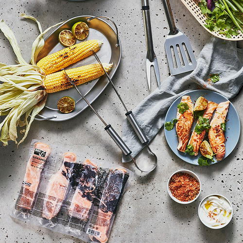 La imagen muestra filetes de salmón de SJÖRAPPORT, maíz asado, pinzas y condimentos en una mesa gris.