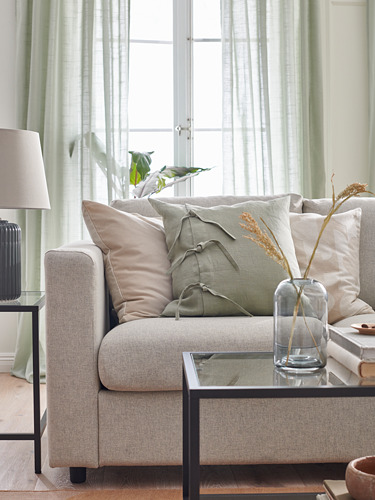 Sala de estar moderna con sofá VIMLE, almohadas neutras y mesa de centro de vidrio con jarrón. Cortinas verdes y lámpara beige.