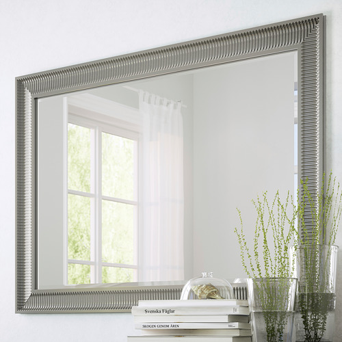 Espejo grande con opciones de suspensión vertical y horizontal, superficie reflectante, con película de seguridad. Se muestra en una ventana reflectante de la pared, junto a plantas y libros.