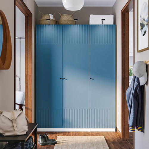 Un gran armario azul PAX domina la habitación, puertas altas de madera, cestas superiores, ganchos para abrigos a la derecha, lámpara en la mesa a la izquierda.